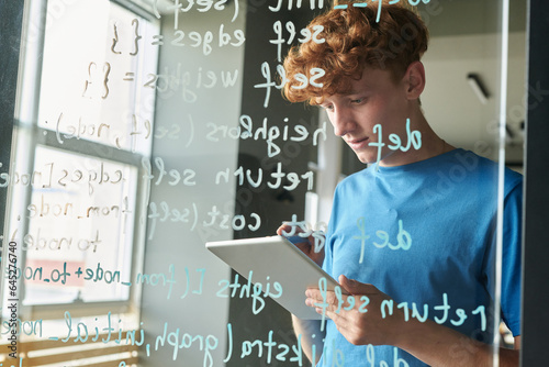 Student using digital tablet while writing computer codes on glass wall at lesson