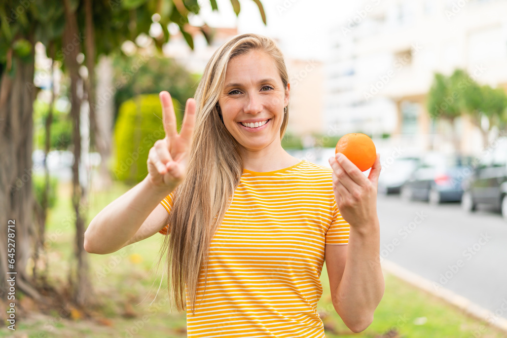 Young blonde woman holding an orange at outdoors smiling and showing victory sign