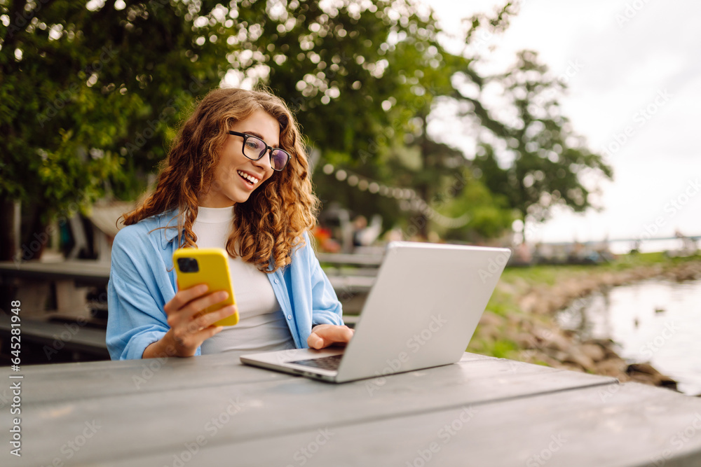 Beautiful female freelancer sitting on a bench at a table in a park outdoors, using a laptop and smartphone. Urban lifestyle. Business, blogging, freelancing. Beautiful female freelancer sitting on a bench at a table in a park outdoors, using a laptop and smartphone. Urban lifestyle. Business, blogging, freelancing.