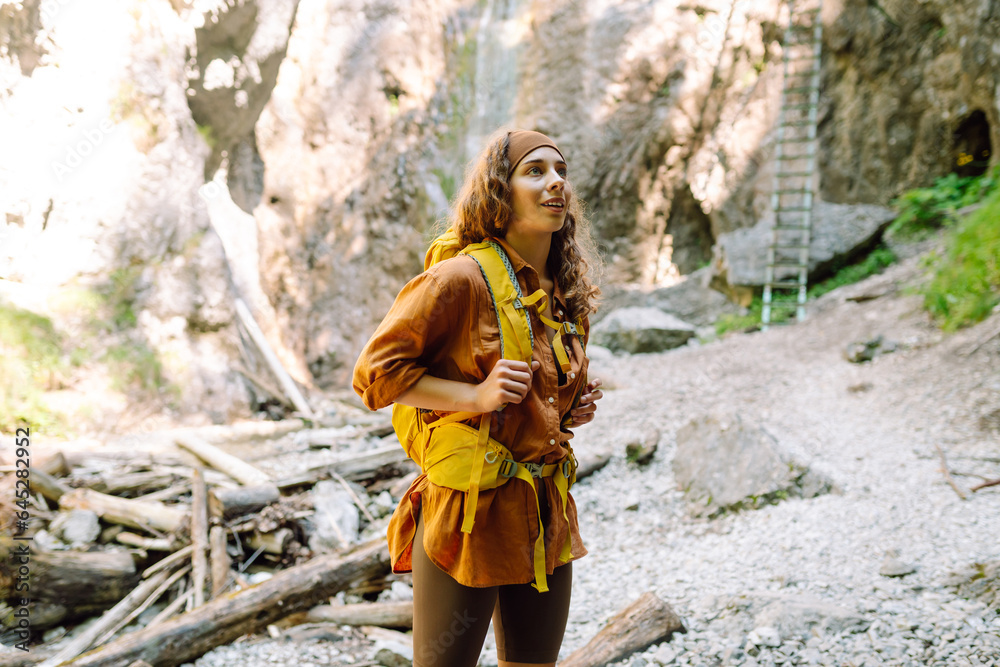 Smiling female traveler with a yellow backpack, dressed in hiking clothes, enjoys a waterfall near a mountain river. Travel, trekking. Nature concept. Smiling female traveler with a yellow backpack, dressed in hiking clothes, enjoys a waterfall near a mountain river. Travel, trekking. Nature concept.