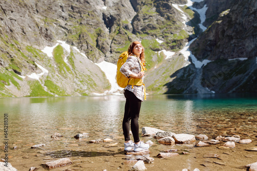 A smiling female traveler with a yellow hiking backpack against the backdrop of a turquoise lake among the mountains. Active lifestyle. Hiking, adventure concept.