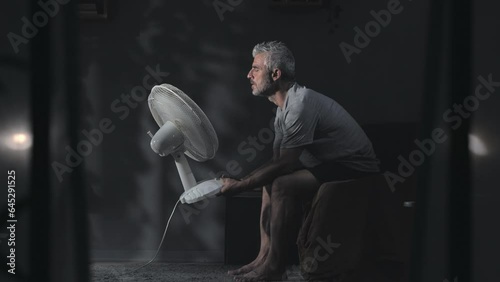 man suffering heat sitting on the bed at night holding fan cooling down,mature male can't sleep at night feeling warm cools with ventilator,summer high temperature