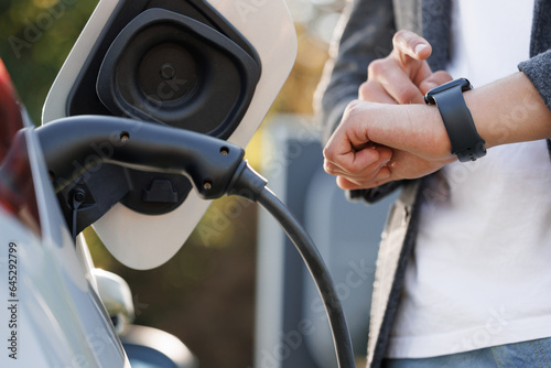 Businessman plugging in charging cable to to electric vehicle. Male hand inserts power connector into EV car and charges batteries, uses smartwatch for activates start charging