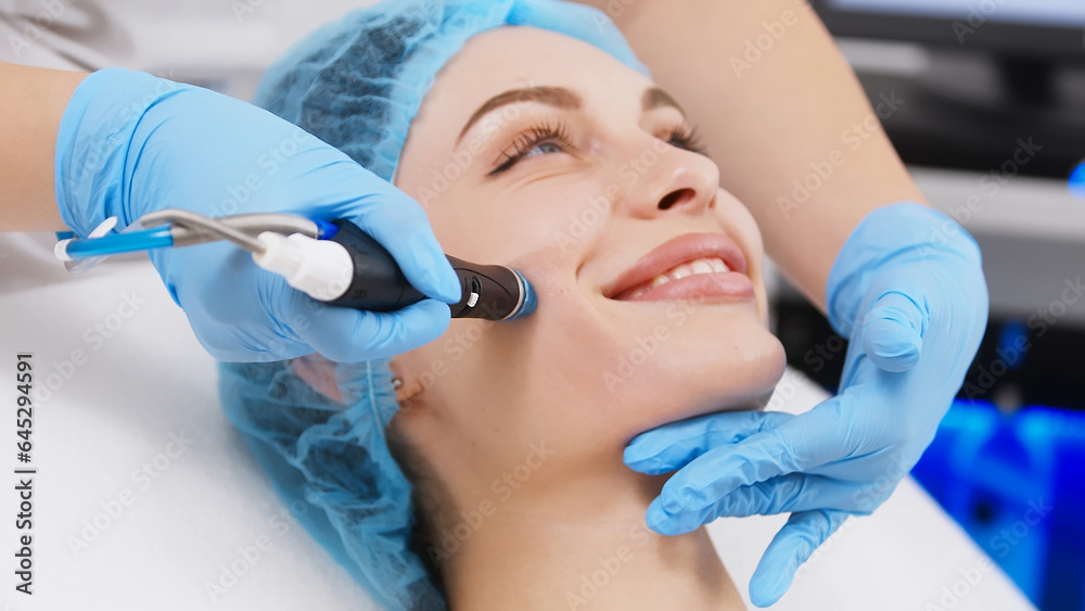 Naklejka premium Cosmetologist doing anti-aging procedure in cosmetology office. Satisfied woman in a disposable hat lies on the sofa and relaxes. Working with the device.