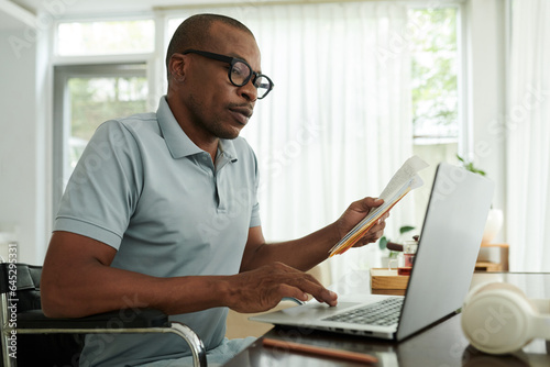 Black man with disability checking his medical documents and filling form on laptop