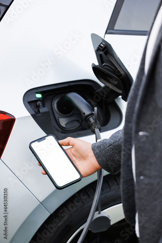 Businessman traveling by electric car having stop at chraging station standing holding smartphone looking at screen copy space for text or product close-up while having vehicle fully charged.