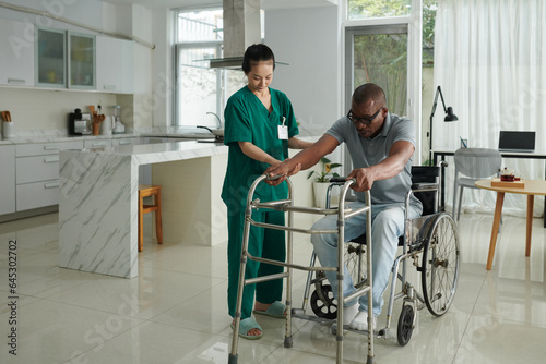 Social worker helping patient to stand up from wheelchair leaning on walkers