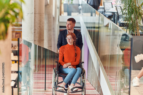 A company director assists his business colleague in a wheelchair, helping her navigate to their startup office, where they work alongside their diverse team of colleagues, emphasizing inclusivity and