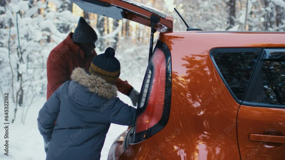 African American man and boy in warm clothes and hats pulling bags out of car trunk and leaving in snowy forest background African American man and boy in warm clothes and hats pulling bags out of car trunk and leaving in snowy forest background