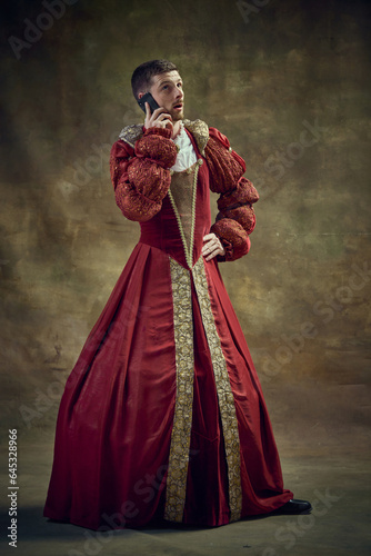Young man in image of medieval royal person wearing female dress, attire and talking on mobile phone on vintage background. Concept of historical retrospectives, fashion, provoking projects, gadgets