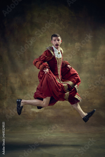 Dramatic apparel. Young man, royal person running on female dress on dark green background. Concept of historical retrospectives, fashion, provoking projects, gender fluidity, masculinity, femininity