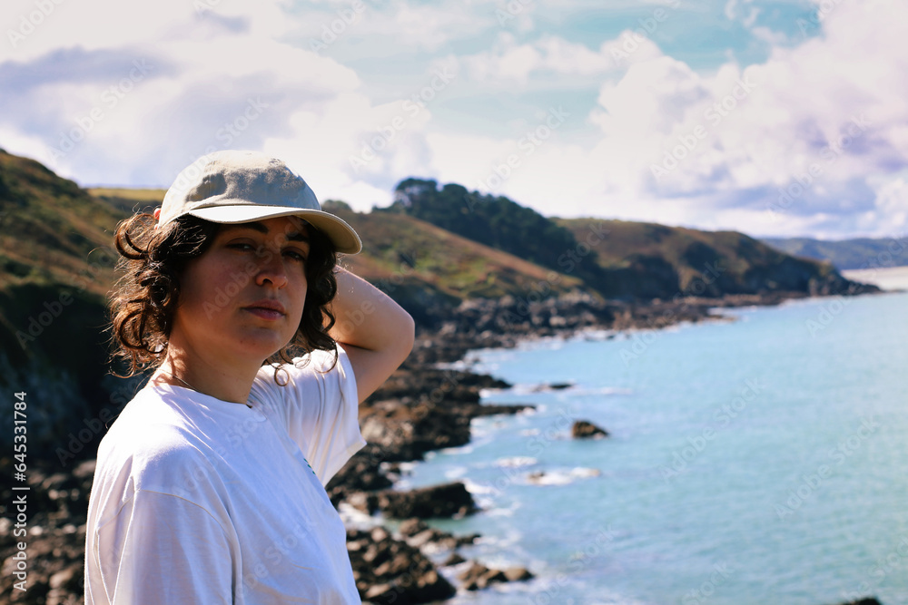 Young woman in cap and white shirt standing on the edge of a cliff by the sea