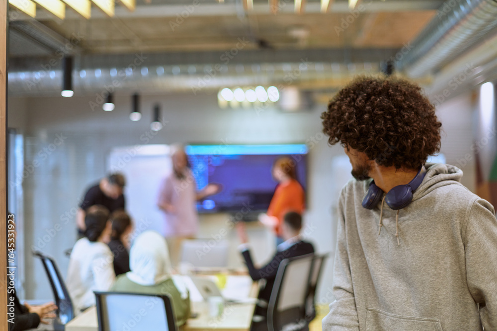 Fototapeta premium In a modern office environment, an African American young entrepreneur with headphones engages in work, while in the background, his dedicated colleagues exemplify teamwork and collaboration