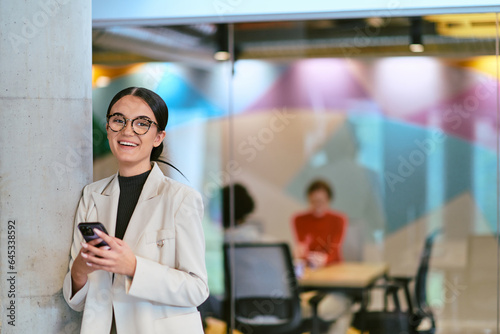 In a modern glass office, a contemporary businesswoman effortlessly multitasks, using her smartphone, while surrounded by colleagues, illustrating the dynamics of corporate life and technology