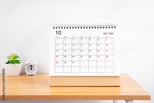 The October Monthly desk calendar for 2023 year and alarm clock on wooden table.