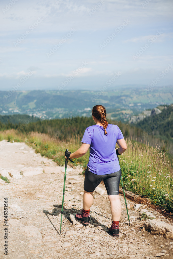 Happy woman hiker with backpack standing on the slope of mountain ridge against mountains. 30s Women holding hiking sticks. Travel and active lifestyle concept, outdoor activities, full body