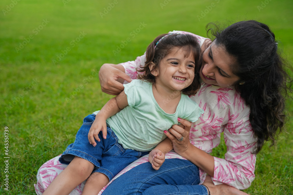 Happy playful young indian mother and cute little girl daughter sitting at summer park or garden having fun together. Happy playful young indian mother and cute little girl daughter sitting at summer park or garden having fun together.