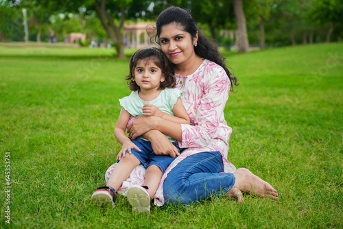 Happy young indian mother and cute little girl daughter sitting at summer park or garden.