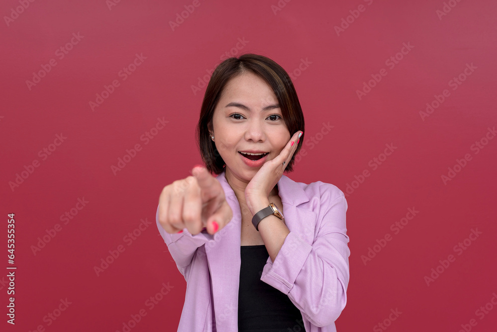 A happily surprised asian woman points in front of the camera. Recognizing someone well known, gushing with excitement. Studio shot with burgundy background. A happily surprised asian woman points in front of the camera. Recognizing someone well known, gushing with excitement. Studio shot with burgundy background.