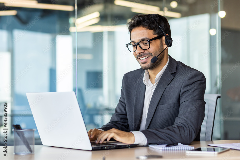 Young successful man at workplace with headset phone typing on laptop keyboard, customer support worker for online buyers inside office, hispanic helping and consulting customers. Young successful man at workplace with headset phone typing on laptop keyboard, customer support worker for online buyers inside office, hispanic helping and consulting customers.