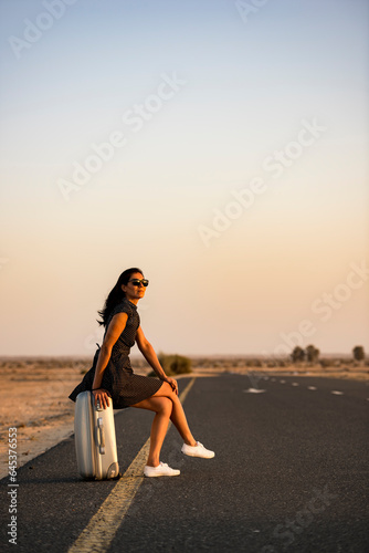 Lonely woman waits on suitcase, deserted road amidst dunes at sunset. Vertical shot with ample copy space.
