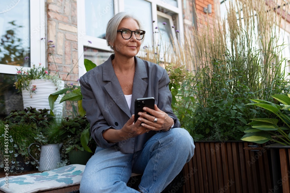 Obraz premium stylish mature woman with gray hair and glasses dressed in a gray jacket chatting in a smartphone on the street