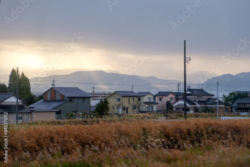 Basking in the radiant glow of the golden sunlight, a picturesque scene unfolds in the Japanese rural landscape. A row of traditional houses stands gracefully amidst the serene countryside,