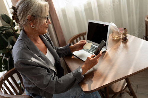 successful well-groomed middle-aged woman with glasses pays for an order using a smartphone and a bank card in a cafe photo