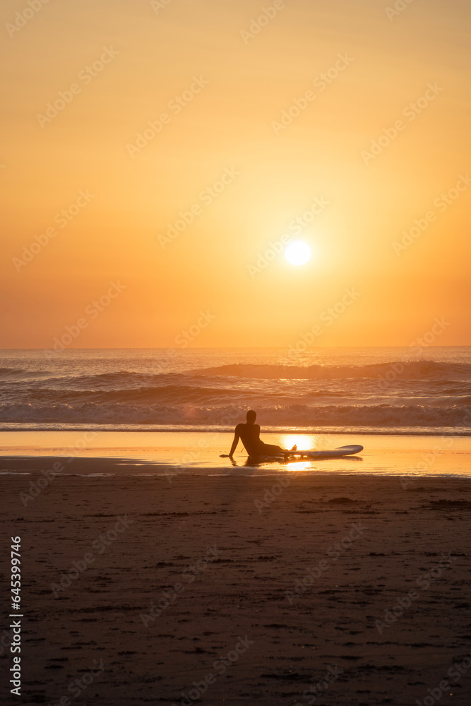 Surfer looks at the golden sun setting on the beach during the sunset