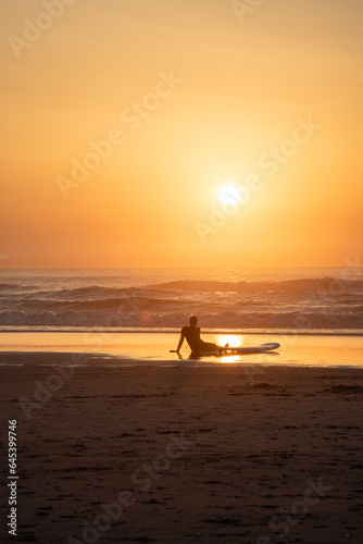 Surfer looks at the golden sun setting on the beach during the sunset