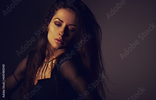 Beautiful woman in black dress posing in fashion necklace on the neck. Long hairstyle on dark shadow background. Toned portrait. Closeup