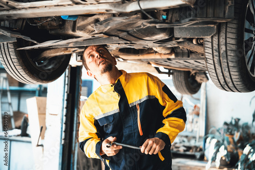 Vehicle mechanic conduct car inspection from beneath lifted vehicle. Automotive service technician in uniform carefully diagnosing and checking car's axles and undercarriage components. Oxus