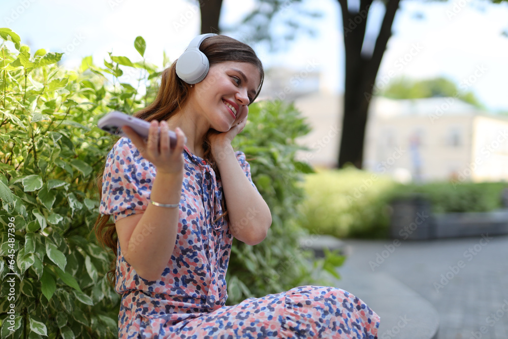 A young woman enjoying her leisure time outdoors in a green park, listening to music through her headphones, and finding relaxation in the soothing melodies.
