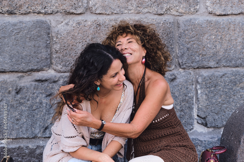 Mother and daughter posing smiling and in complicity sitting in front of a stone wall. Mother and daughter posing smiling and in complicity sitting in front of a stone wall.
