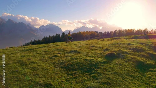 AERIAL: Stunning alpine views at the hilltop enjoyed by a woman with her dog. Relaxed sporty lady sits on electric bike and admires last rays of autumn sun illuminating beautiful mountain landscape. photo