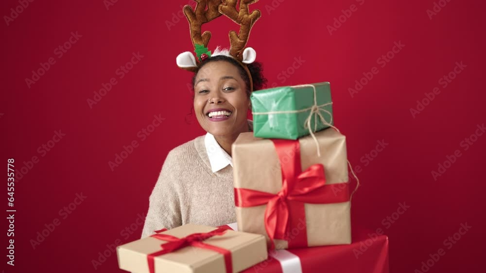 African american woman holding christmas gifts wearing reindeer ears over isolated red background African american woman holding christmas gifts wearing reindeer ears over isolated red background