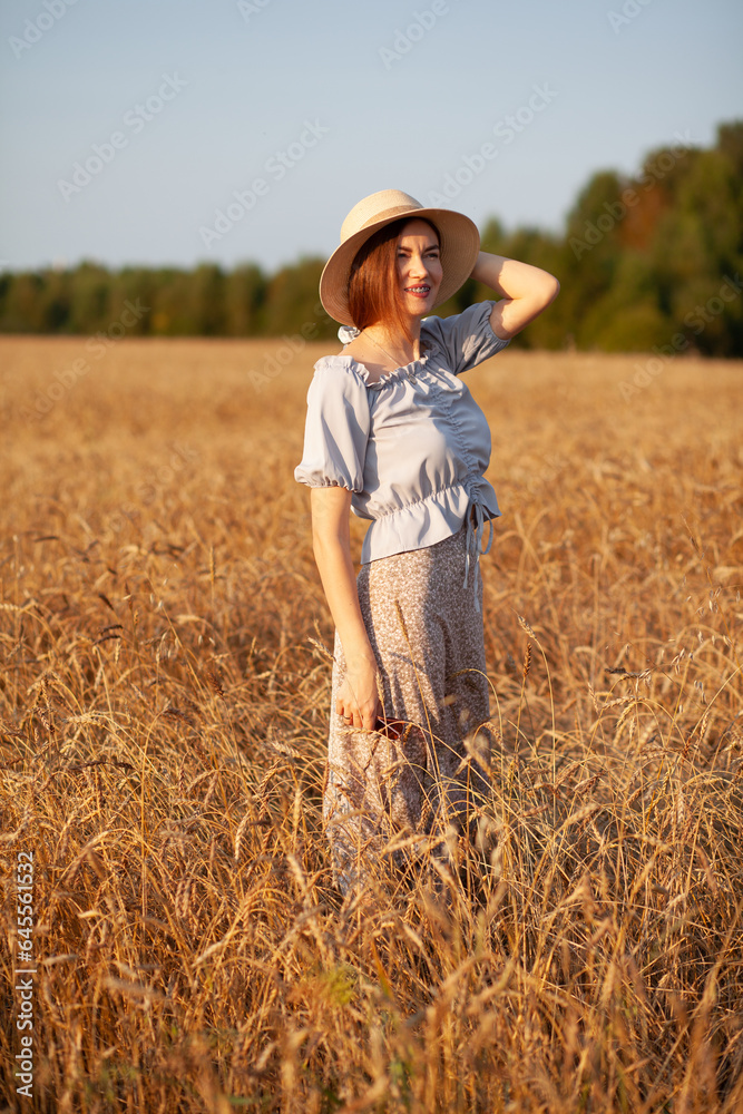 A young beautiful girl with braces on her teeth and long hair poses in a wheat field in the summer at sunset. The girl holds a hat in her hand against the background of a wheat field.