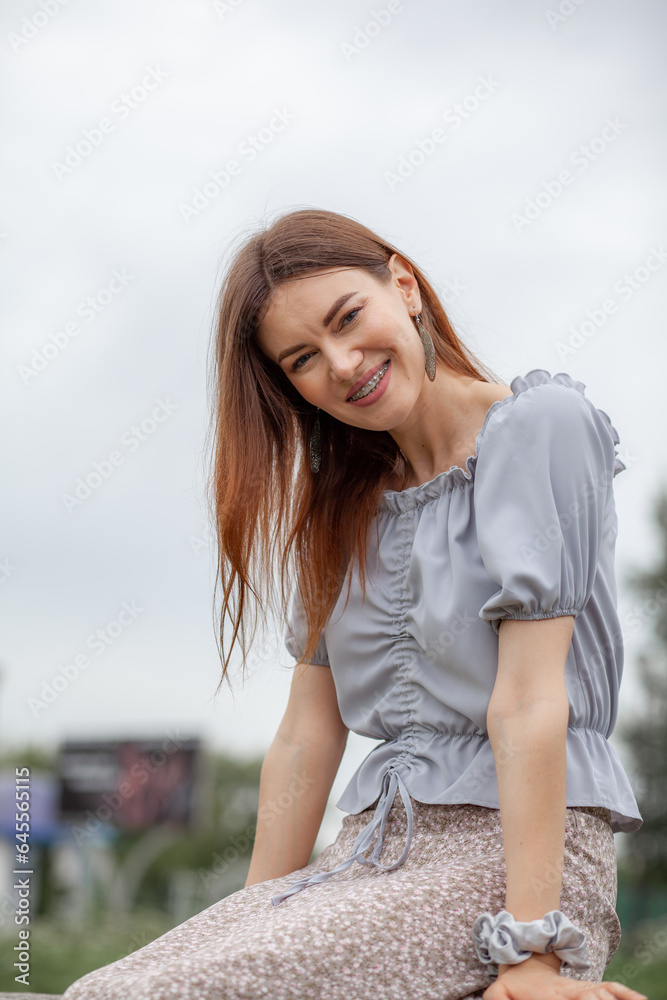 Smile of a young and beautiful girl with braces on her white teeth. A girl with long hair and a dress poses in the park. Straightening crooked teeth using a braces system.