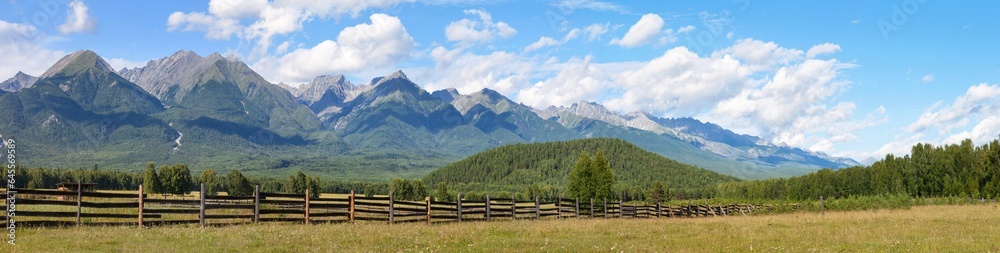 Panoramic view of beautiful mountain landscape with hay meadow and rocky ridge of mountains against blue sky with clouds on sunny August day.  Eastern Sayan Mountains, Tunka valley, Buryatia