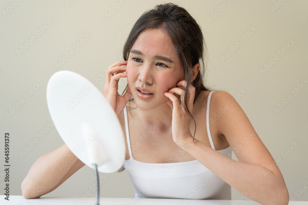 Asian young woman looking face skin in the mirror have a red rash on her face from cosmetic allergy