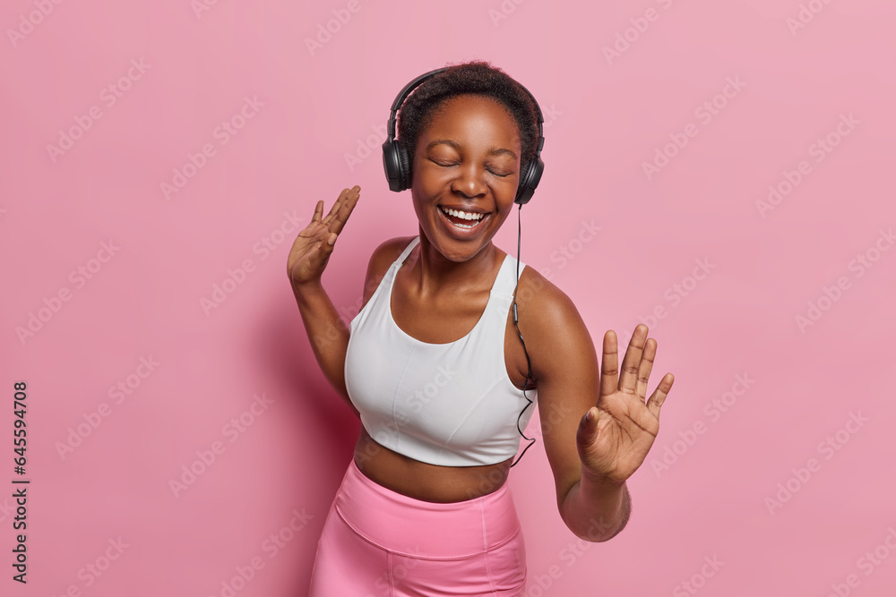 Positive energetic sporty woman moves with rhythm of music wears stereo headphones on ears wears sportswear keeps eyes closed isolated over pink background. People fun and lifestyle concept. Positive energetic sporty woman moves with rhythm of music wears stereo headphones on ears wears sportswear keeps eyes closed isolated over pink background. People fun and lifestyle concept.