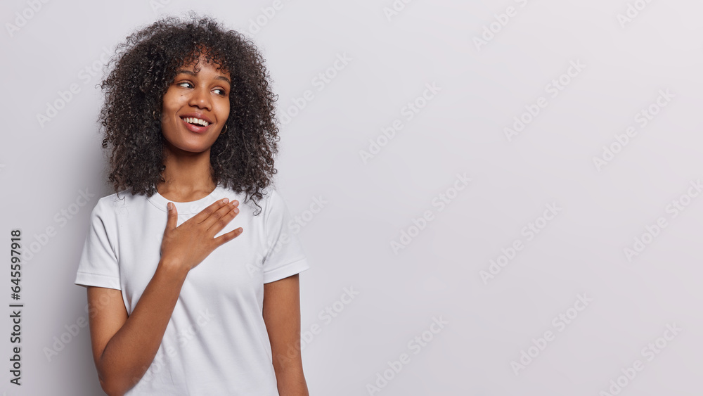 Horizontal shot of beautiful curly haired woman keeps hand on chest feels thankful and delighted smiles pleasantly dressed in casual t shirt isolated over white background empty space for your advert