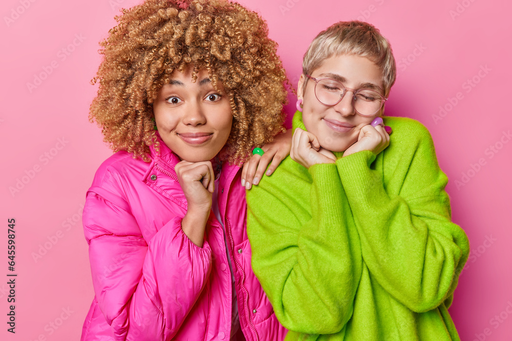 Positive emotions concept. Indoor waist up of happy smiling African american and European ladies standing close to each other isolated on pink background wearing casual clothes keeping hands raised