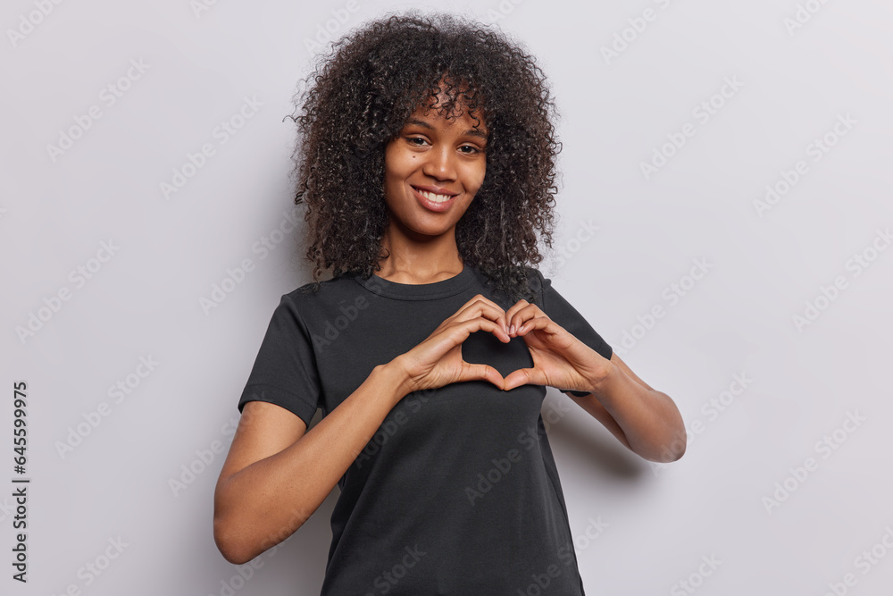 Horizontal shot of friendly black woman with curly hair makes love gesture expresses kindness dressed in casual black t shirt isolated over white background. Romantic female model shows heart sign - obrazy, fototapety, plakaty 
