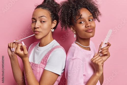 Beauty procedures. Indoor shot of two pretty young glad African american females wearing casual clothes standing in centre back to back on pink background doing manicure trying to look at each other
