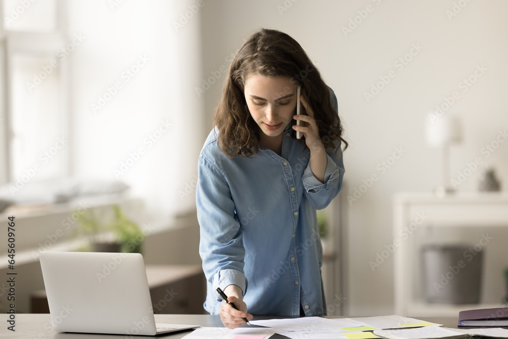 Busy young gen Z business woman talking on mobile phone at home office work table, checking paper reports, writing notes, discussing project plan, startup strategy on cellphone Busy young gen Z business woman talking on mobile phone at home office work table, checking paper reports, writing notes, discussing project plan, startup strategy on cellphone