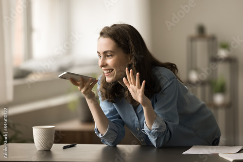 Happy inspired gen Z young freelancer girl discussing creative idea for business project in mobile phone, talking on speaker, holding smartphone at mouth, smiling, laughing, speaking