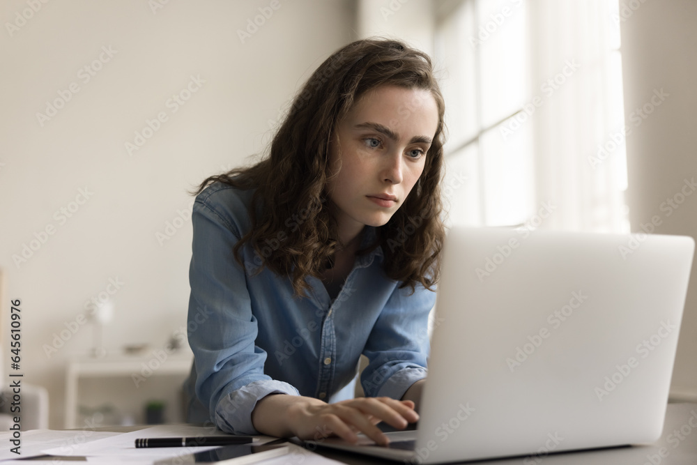 Serious young gen Z business woman typing at laptop, bending over workplace table engaged in process, looking at display, writing down creative idea for project Serious young gen Z business woman typing at laptop, bending over workplace table engaged in process, looking at display, writing down creative idea for project