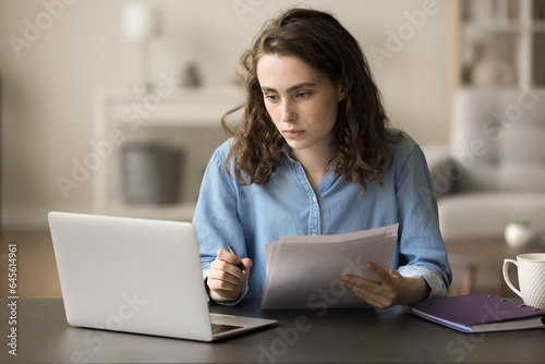 Serious busy young freelancer girl checking financial papers, legal documents at work table, using at laptop, looking at display, reviewing contract, agreement, consulting online paperwork service