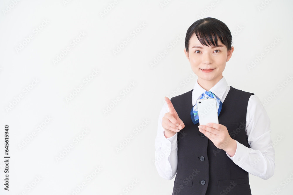 スマートフォンを操作するベストにリボンタイの制服を着た、受付業務、事務業務の女性 スマートフォンを操作するベストにリボンタイの制服を着た、受付業務、事務業務の女性